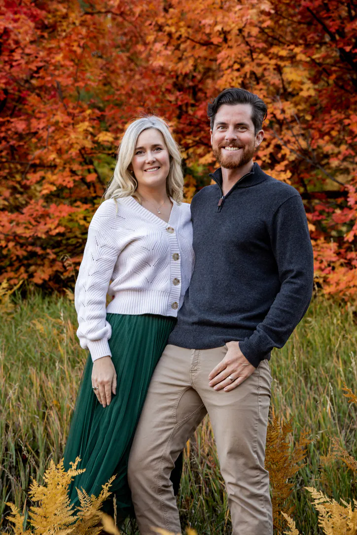 Jim et Ashley, côte à côte dans les hautes herbes d'automne, devant des arbres rouges et orange.